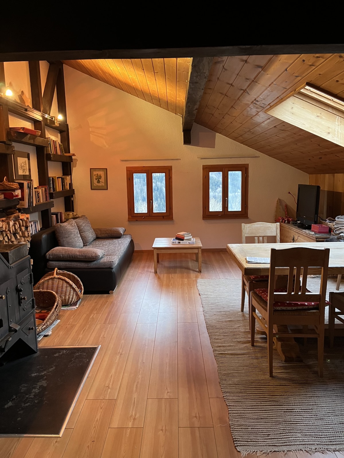 A cozy living area features a sofa positioned near windows that provide natural light. A wooden dining table is accompanied by chairs, and bookshelves line the walls. A wood stove is visible, contributing to the interior's inviting ambiance.