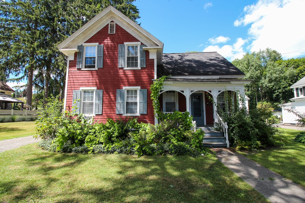 A two-story red house features a welcoming front porch with white trim. Lush greenery surrounds the entrance, along with an expansive lawn. Large windows allow natural light to illuminate the home, contributing to its inviting appearance.