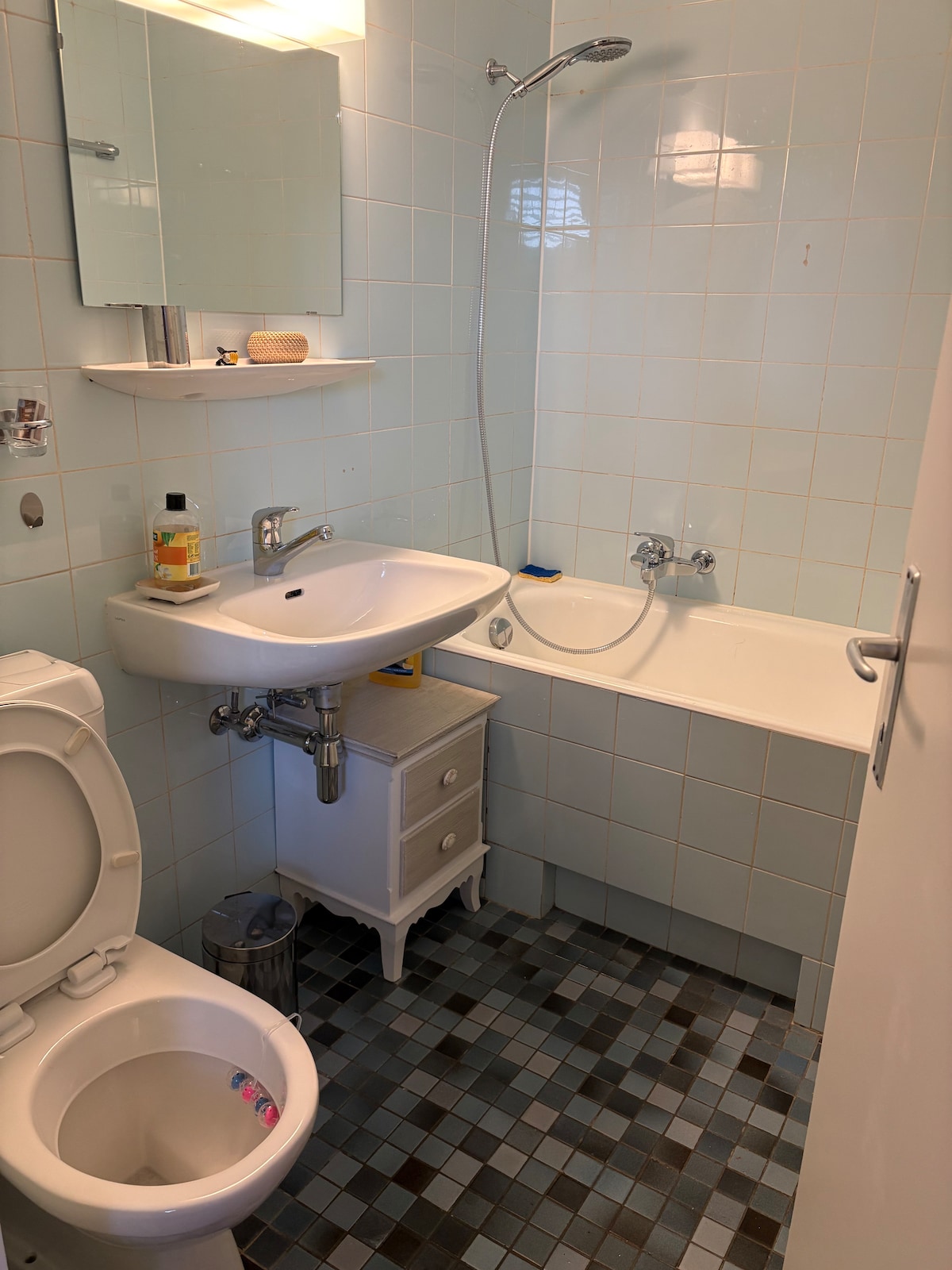 A functional bathroom is presented, featuring a sink and a bathtub with a shower attachment. Light blue tiles cover the walls, while a small white cabinet is positioned beside the sink. A modest toiletries shelf is mounted above the sink, and a waste bin is placed in the corner.