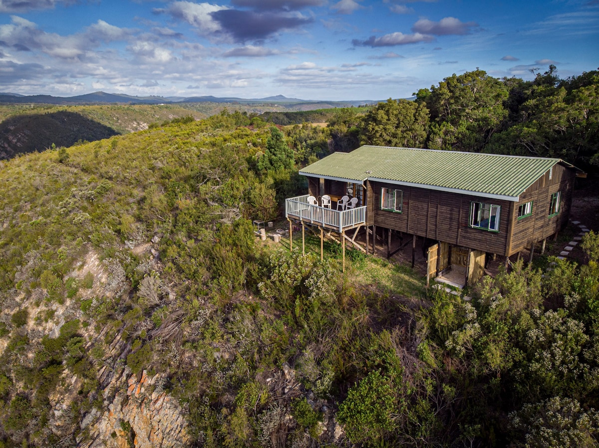 A rustic cottage is situated on the edge of a lush ravine, surrounded by greenery and showcasing expansive views of the Outeniqua Mountains. A spacious deck with outdoor seating extends from the cottage, offering a serene space for relaxation.