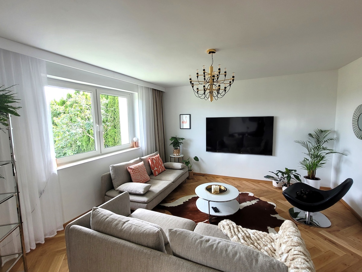 A spacious living room features two comfortable sofas and a stylish armchair, arranged around a central coffee table. A large flat-screen television is mounted on the wall, while natural light enters through two large windows adorned with light curtains. Potted plants add freshness to the space.