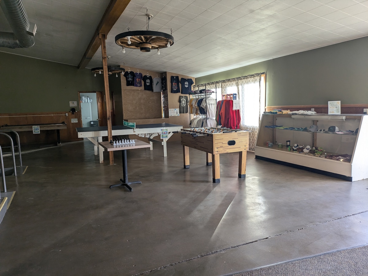 A spacious common area is presented with polished concrete floors and green walls. A foosball table and a table for games are situated in the center, while a display cabinet with various items stands to the right. Natural light enters through a nearby window, illuminating the space.