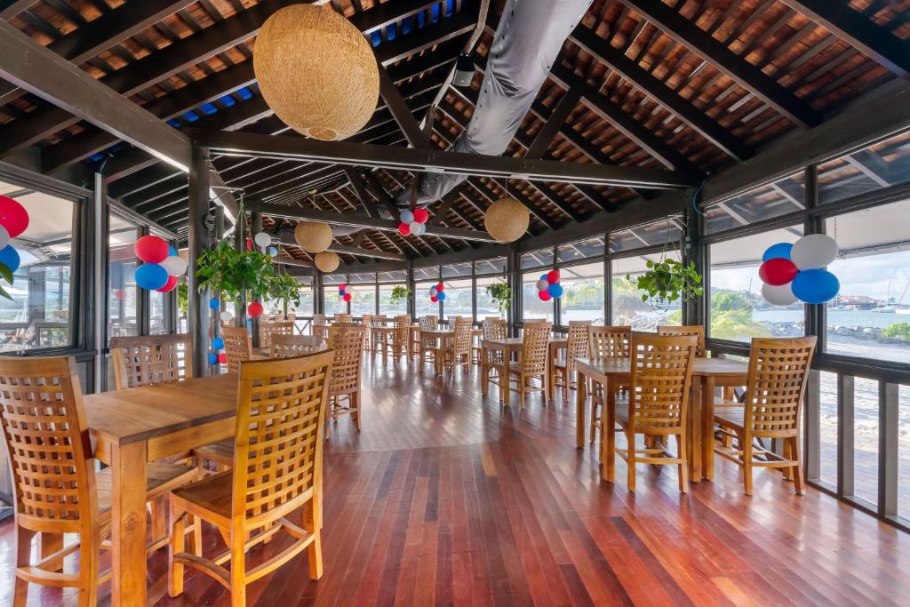 A spacious dining area features wooden flooring and tall ceilings supported by beams. Tables are arranged for dining, each adorned with woven chairs. Decorative elements include hanging light fixtures and colorful balloons in red, white, and blue, adding a festive touch to the atmosphere.