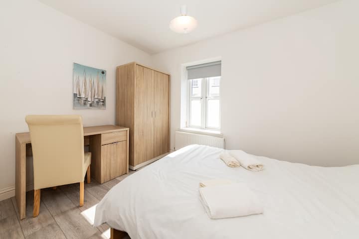 Un dormitorio luminoso y minimalista con una cama doble, sábanas blancas, dos toallas dobladas, una mesita de noche de madera con una lámpara, un armario, una ventana con persianas y un mapa colgado sobre la cama.