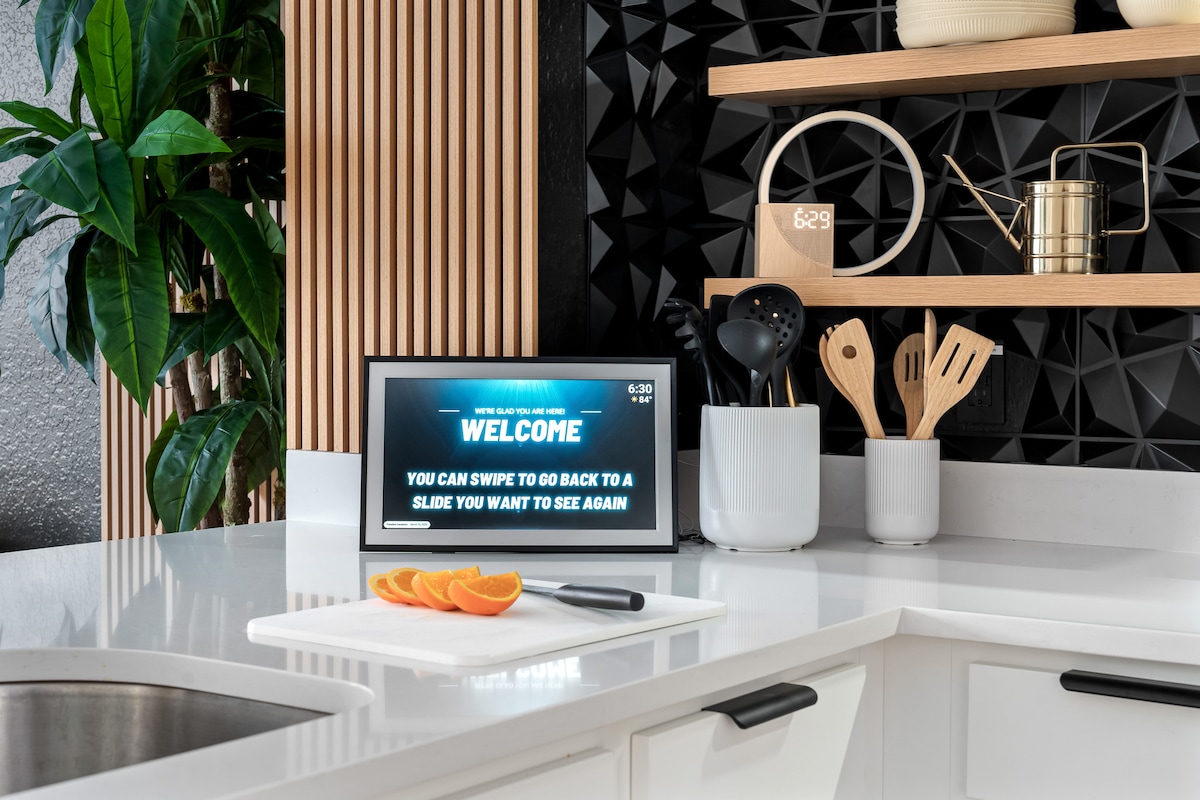 A modern kitchen space features clean white cabinetry and a sleek countertop. An interactive display is positioned on a counter, welcoming guests. Kitchen utensils and a cutting board with fresh oranges are arranged neatly beside a potted plant, adding a touch of greenery.