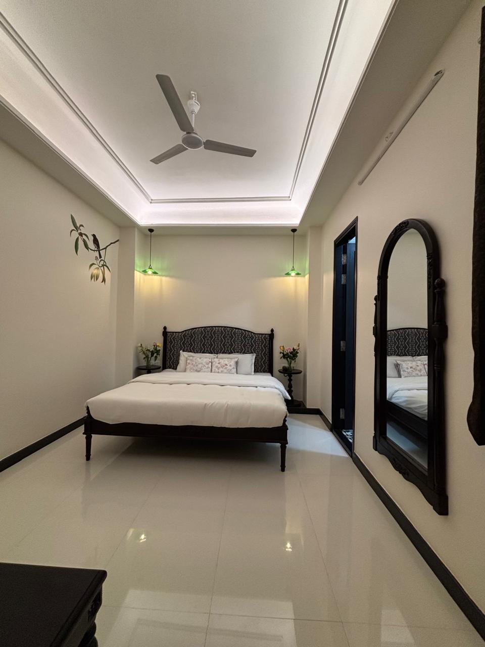 A well-appointed room features a spacious bed with a patterned headboard. Soft lighting is provided by green sconces on either side. A tall mirror and floral accents contribute to the room's decor, while glossy tiles create a bright and clean atmosphere.
