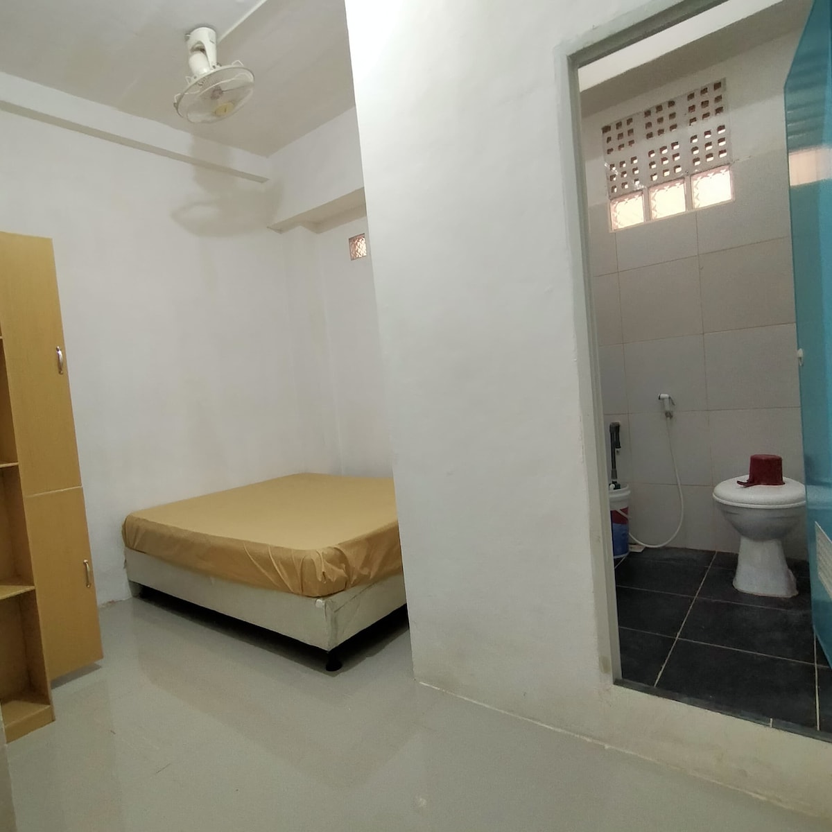A simple sleeping area is shown with a spacious bed featuring a beige cover. Built-in shelving is visible along one wall, complementing the neutral tones of the room. An adjoining bathroom is partially seen, equipped with a shower and toilet.