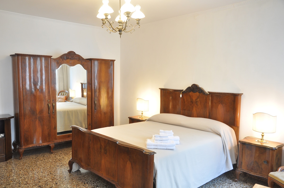 A spacious bedroom features a large wooden bed flanked by matching nightstands with lamps. A vintage wardrobe adds character, reflecting the room's warm tones. Natural light fills the space, evident from the mirror opposite the bed, enhancing the tranquility of the environment.
