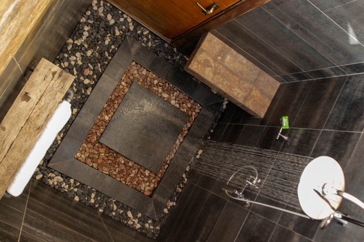An outdoor shower space is presented, featuring a smooth stone floor bordered by various pebbles. A wooden bench offers seating alongside the showerhead, surrounded by sleek, dark tiles that evoke a natural ambiance.