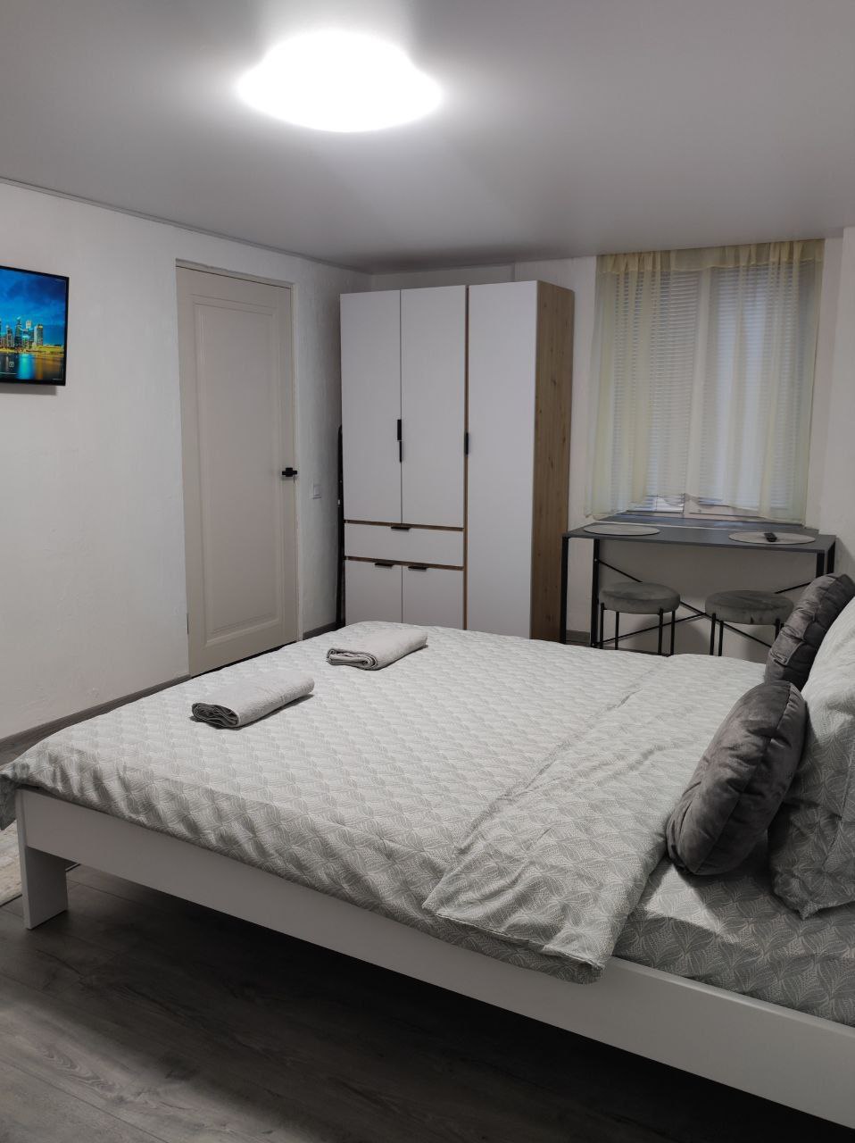 A welcoming bedroom features a spacious bed dressed in light grey linens. A dresser stands against the wall, and a small dining table with two chairs is positioned near a window adorned with sheer curtains. Two rolled towels are neatly placed on the bed.