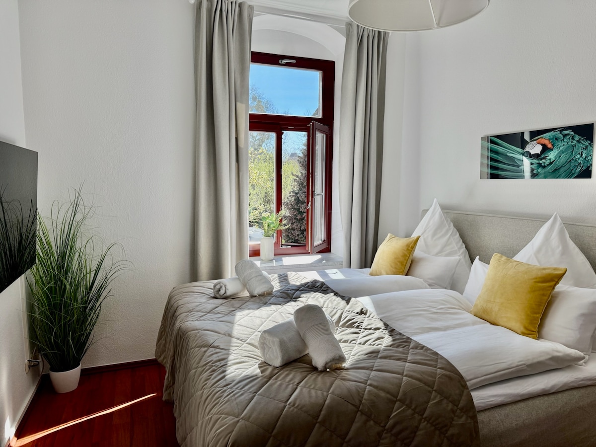 A cozy sleeping area features a spacious bed with neutral bedding and decorative yellow cushions. Natural light floods the room through large windows, partially open to reveal greenery outside. A plant adds a touch of nature, while soft curtains provide a gentle contrast.