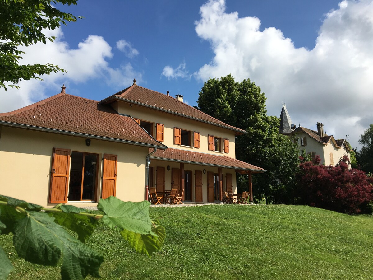 The exterior of a two-story house features a light-colored facade and wooden shutters. A spacious lawn is visible in the foreground, with several chairs arranged on a patio, surrounded by trees. The sky is partly cloudy, providing a balanced light across the scene.
