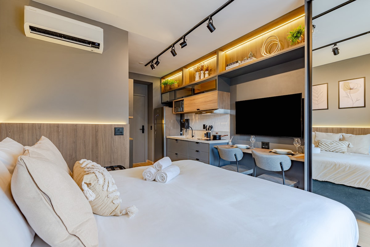 A modern studio apartment features a spacious bed with plush pillows and neatly arranged towels. A well-equipped kitchenette and dining area are visible, alongside a wall-mounted smart TV. Ambient lighting highlights the stylish decor, contributing to a functional and welcoming design.