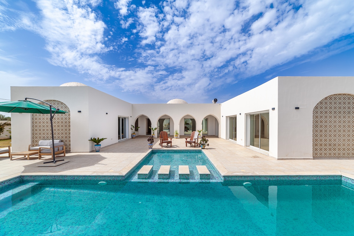 A modern villa exterior is presented, featuring a clear blue sky and unique architectural arches. A private pool is visible, accompanied by lounge chairs and a shaded area. Large glass doors provide access to the inviting interior, creating an open and airy atmosphere.