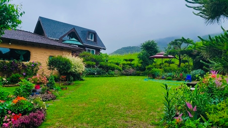 A well-maintained garden bursts with colorful flowers and lush greenery, surrounding a charming two-story house. A covered gazebo is visible amidst the vibrant landscaping, with gentle hills in the background that complement the serene outdoor atmosphere.
