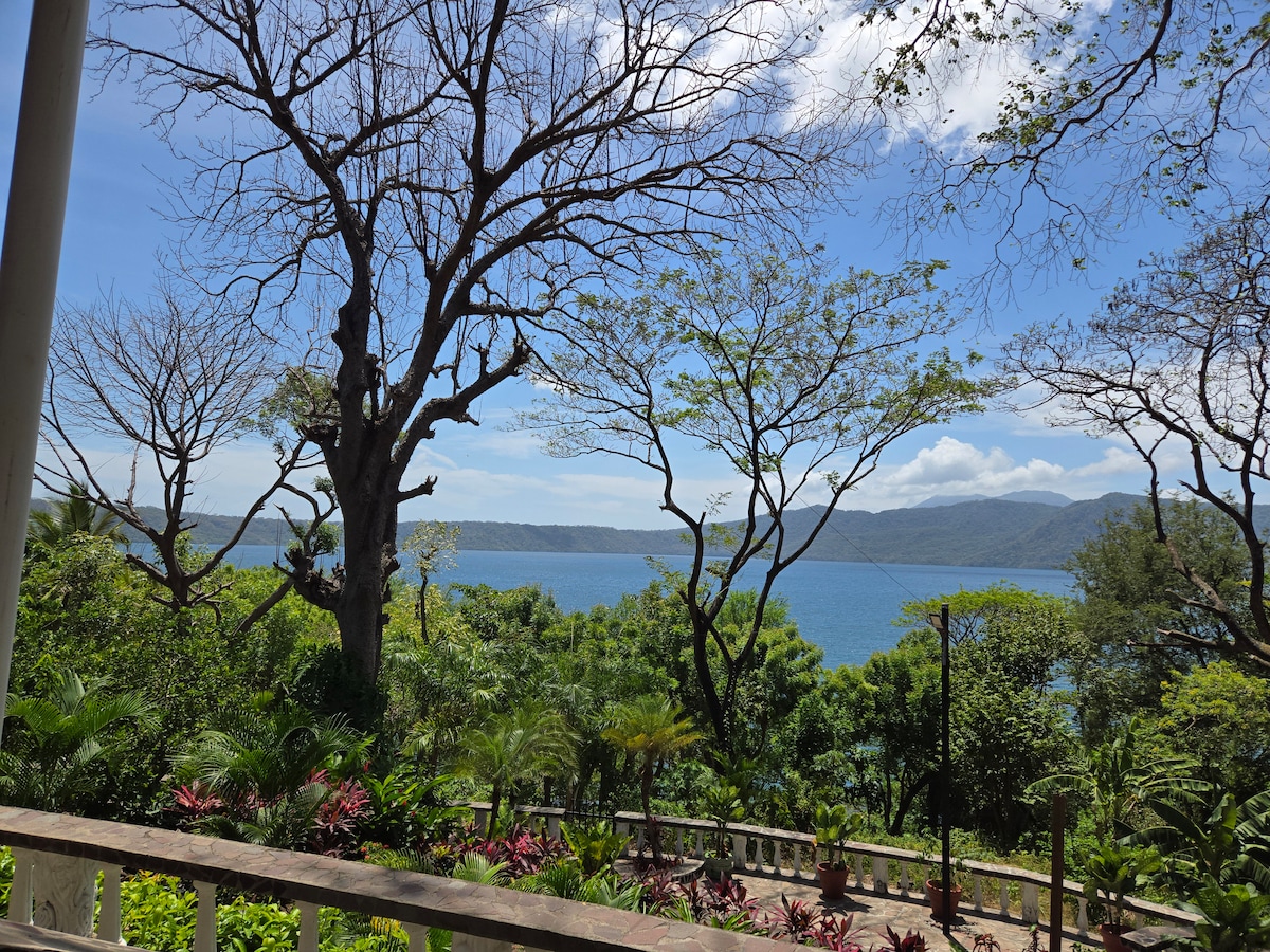 A serene landscape is presented, featuring a calm body of water framed by lush greenery and trees. The blue sky and distant mountains are visible, creating a peaceful, natural setting.