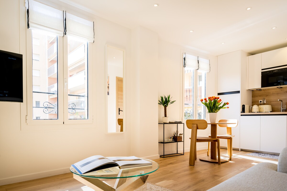 A bright living space features large windows allowing natural light to fill the room. A simple glass coffee table is positioned in the foreground, while a wooden dining table with chairs is seen in the background, complemented by a modern kitchen setup.