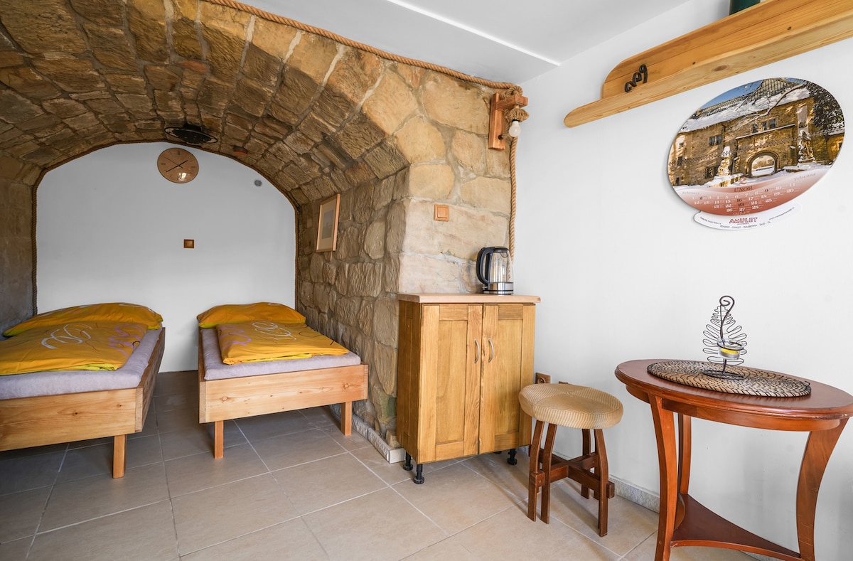 Two twin beds with yellow linens are positioned in an alcove with a stone arched ceiling. A wooden cabinet holds a kettle, while a small round table with a decorative item is placed nearby. Natural light enters from a wall-mounted clock.
