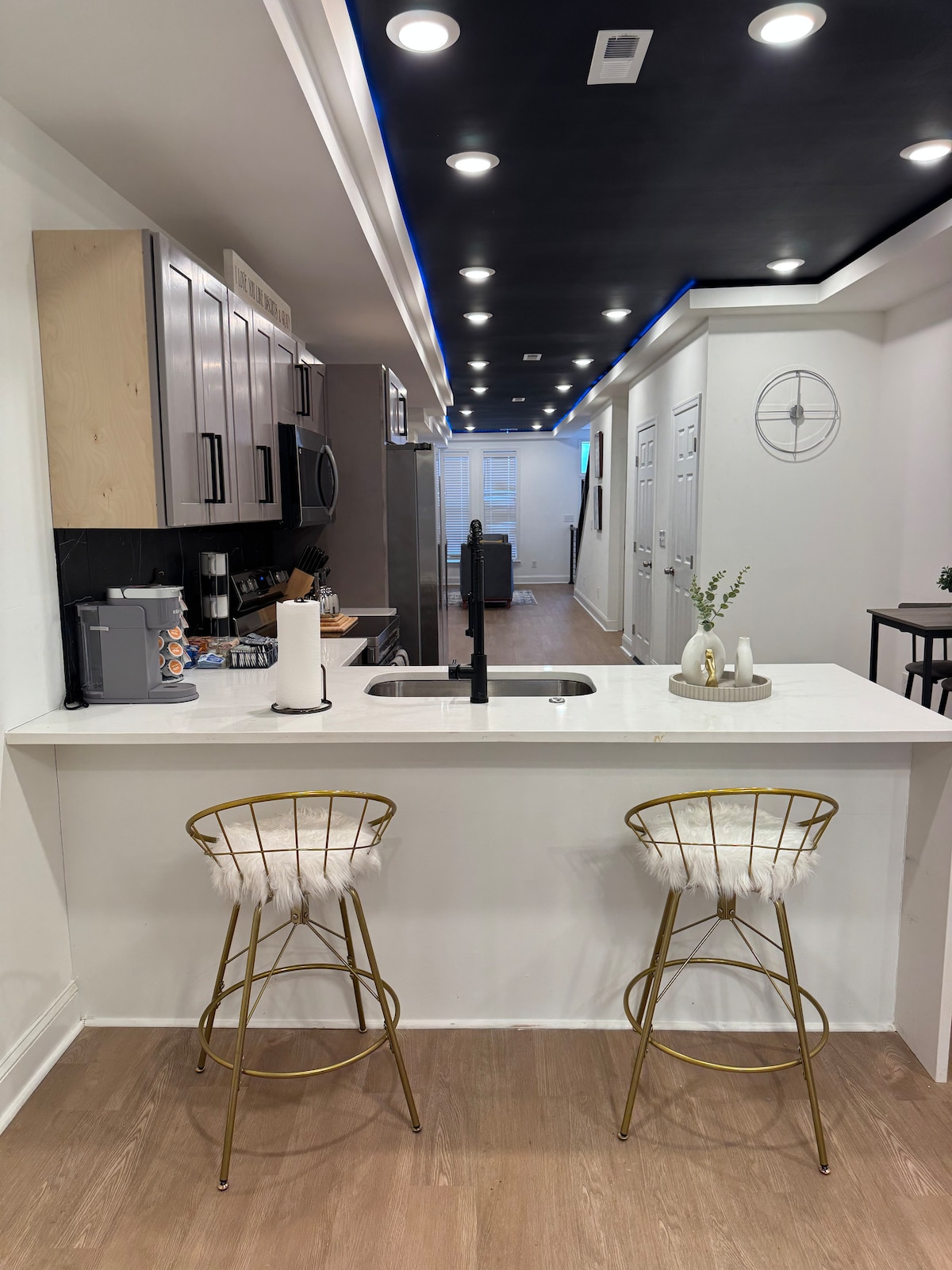 A modern kitchen area is highlighted by a white countertop with a sleek black faucet. Two gold bar stools, adorned with soft white padding, are positioned at the counter. The background reveals an open layout leading to the living space, accented by LED lighting in the ceiling.