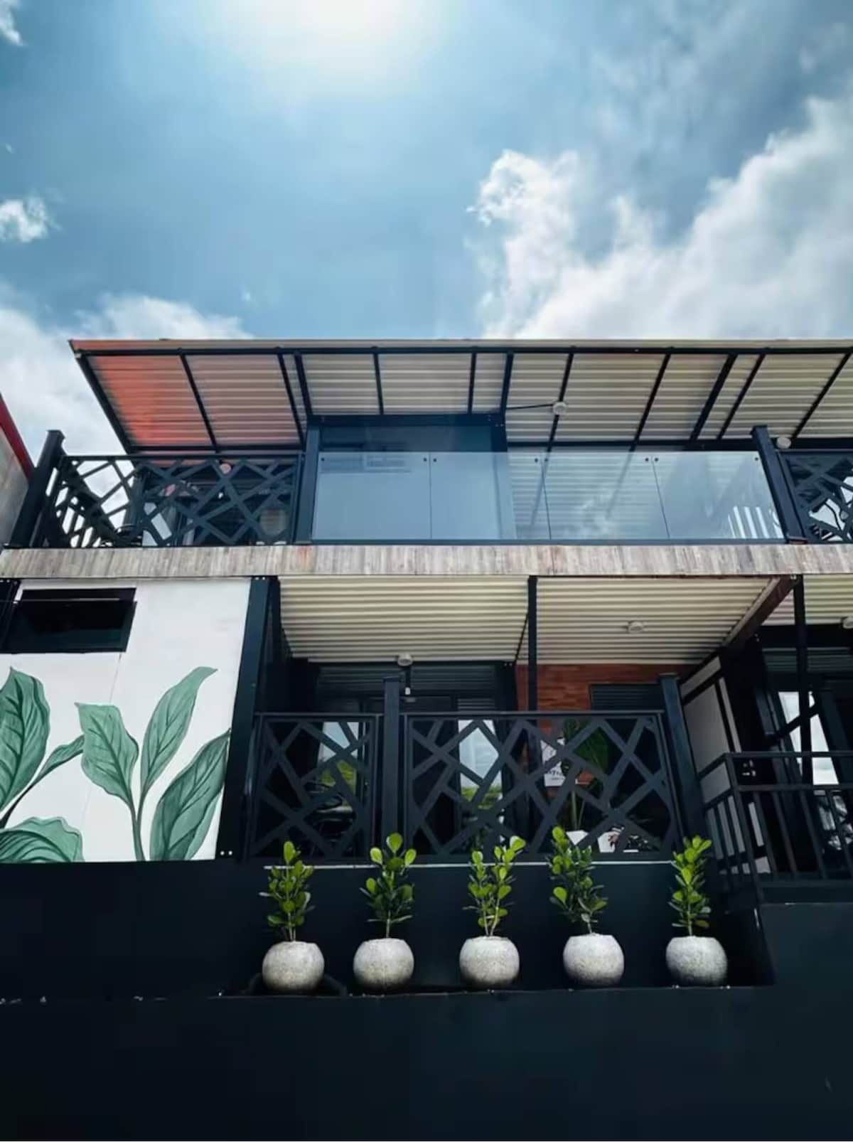 A modern two-story building is showcased against a blue sky, featuring large glass panels and a balcony. Decorative plants in round pots line the ground level, with green leaf motifs visible on the exterior wall.