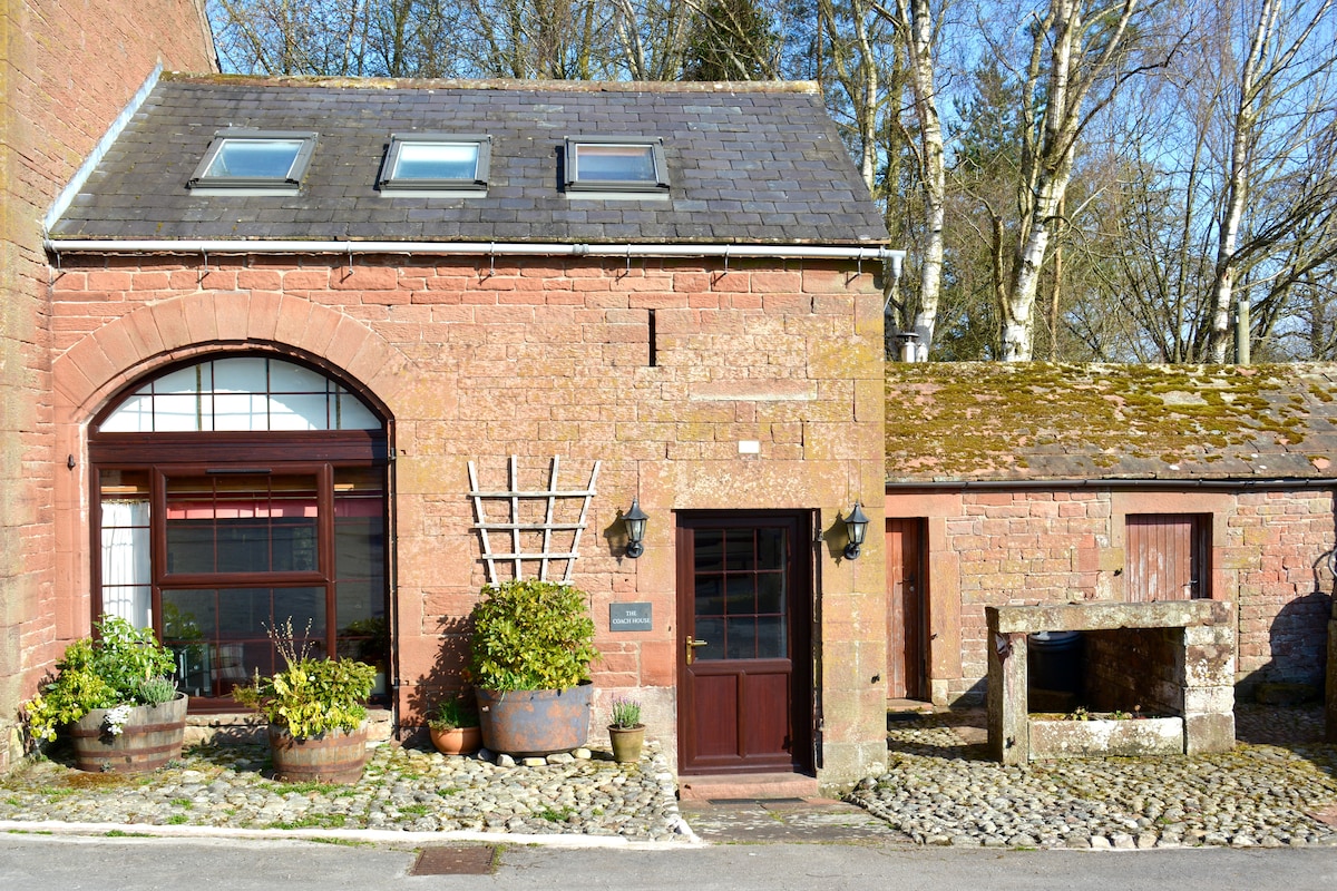 The stone exterior displays a combination of arched and rectangular windows, enhancing the building's character. A wooden door, framed by decorative planters, offers an inviting entrance. The surrounding area features cobbled pathways and greenery, suggesting a serene rural setting.