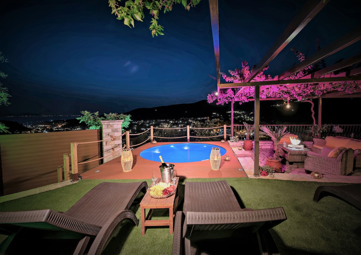A tranquil outdoor area features a round swimming pool illuminated by soft lighting, surrounded by sun loungers on lush green grass. A seating arrangement, framed by a wooden pergola, showcases cozy cushions, while panoramic views of the village and night sky enhance the serene ambiance.