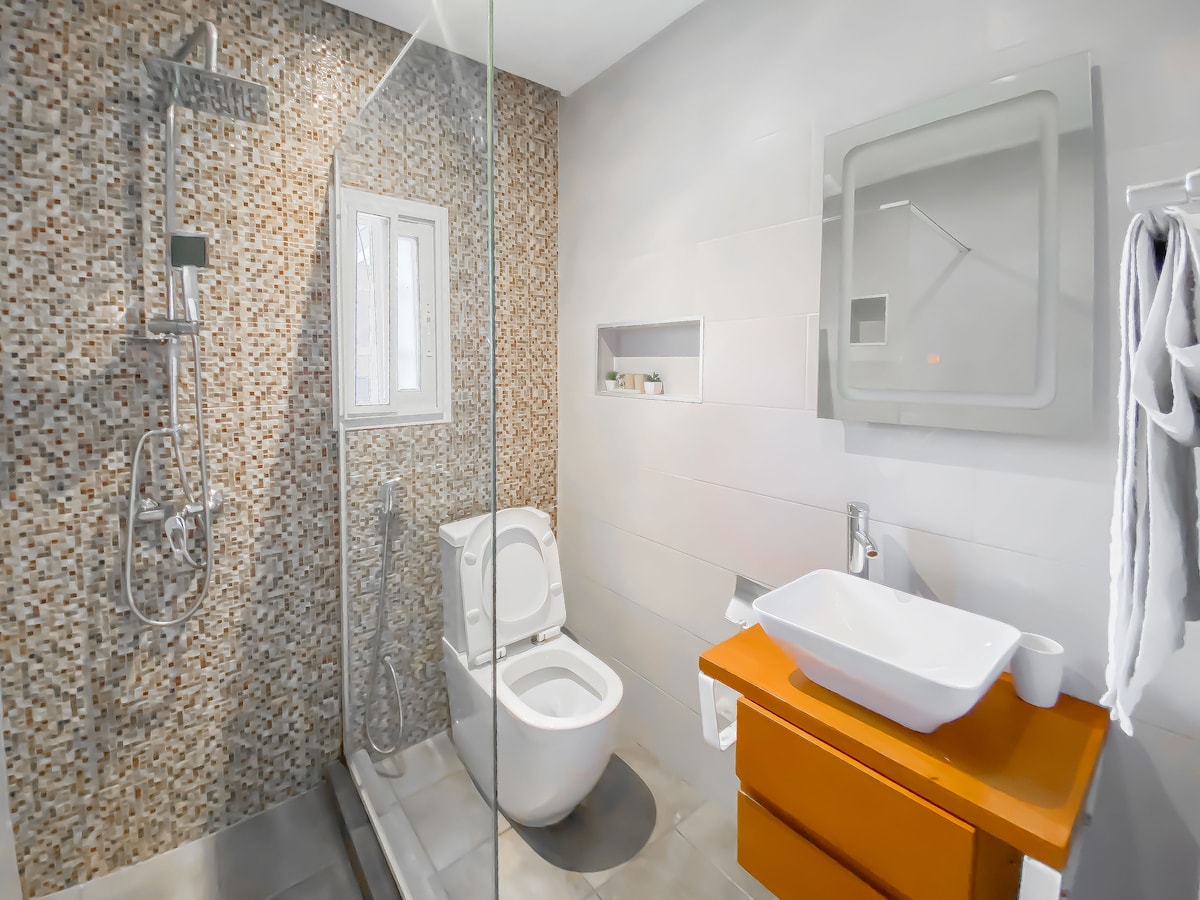 A modern bathroom features a walk-in shower with a contemporary showerhead against a textured mosaic wall. A white toilet is positioned alongside a sleek orange vanity with an integrated sink. Natural light filters through a small window, enhancing the clean and functional design.