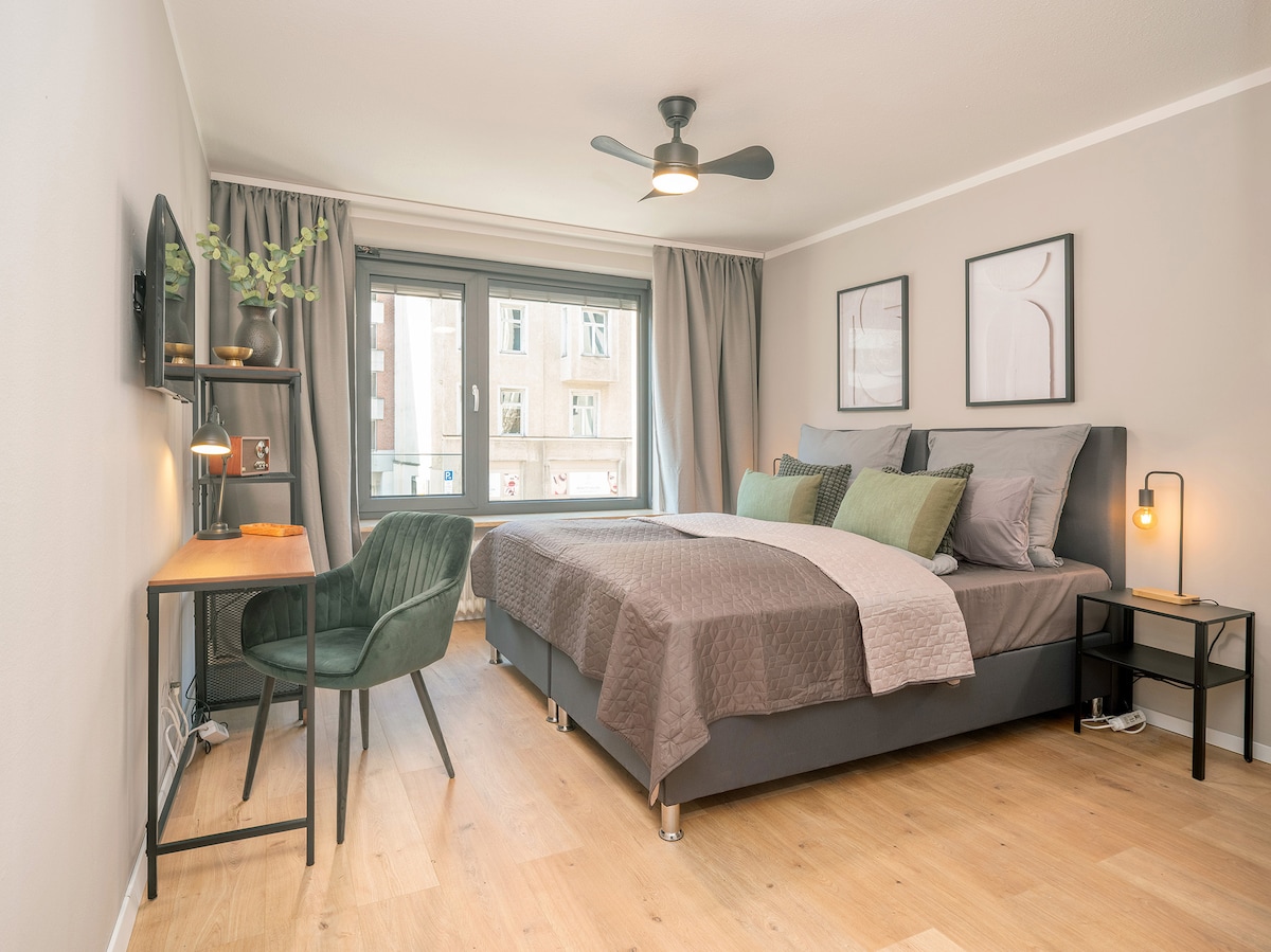 A modern room features a spacious bed with neutral bedding and decorative cushions. A small desk with a green chair is positioned near a window adorned with light curtains. A wall-mounted TV and framed art pieces are displayed, enhancing the room's contemporary design.