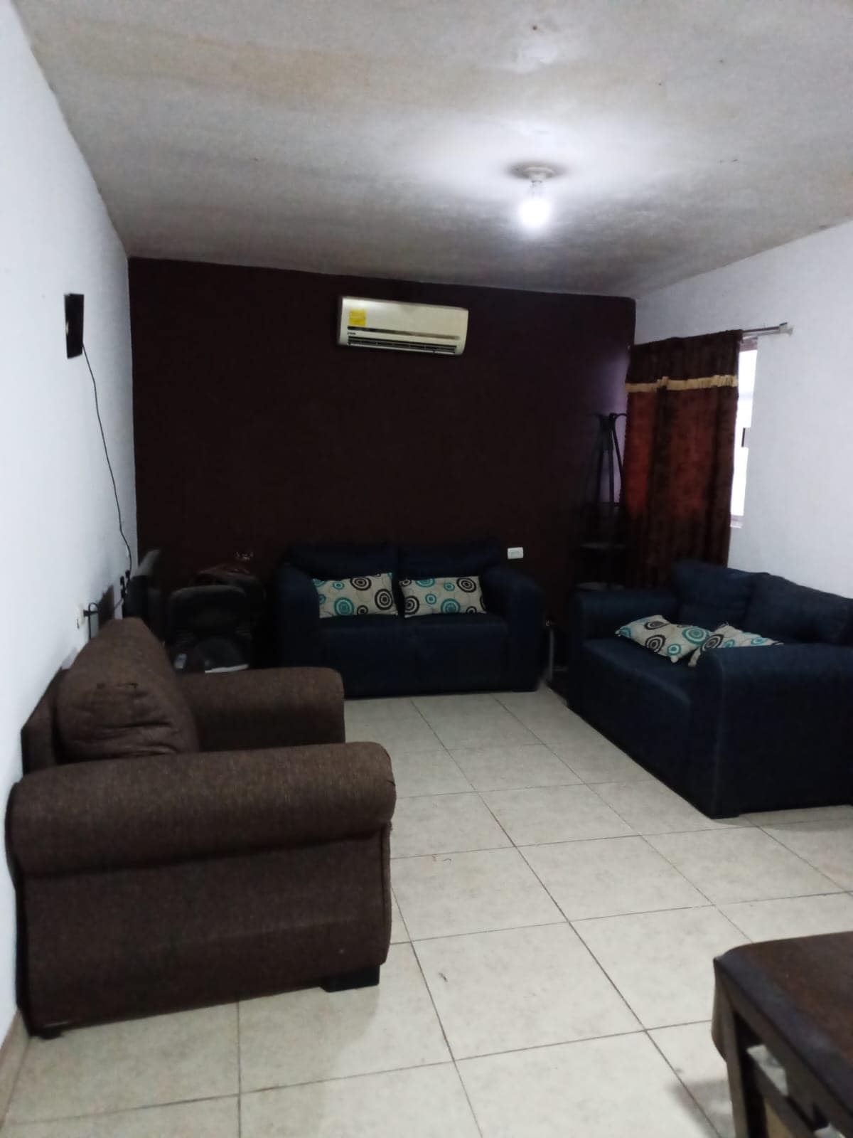 A cozy living area features two upholstered sofas in contrasting colors, with decorative cushions on both. A wall-mounted air conditioning unit is visible above, and natural light enters through a window dressed with dark curtains. Neutral tile flooring complements the overall space.