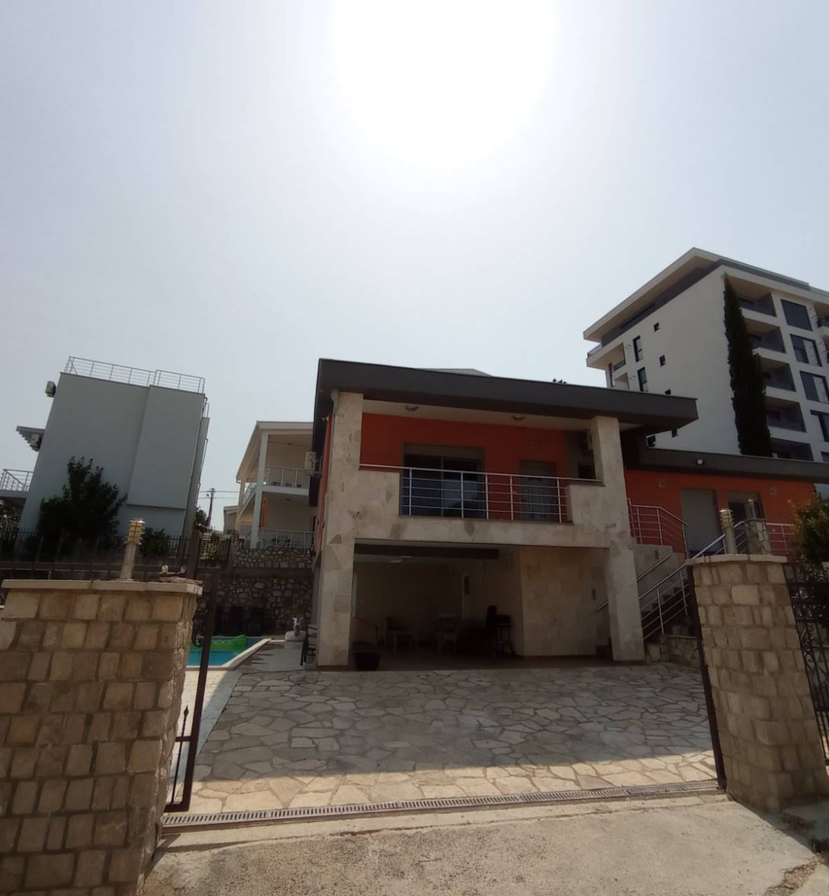 The villa features a two-story structure with a modern design, showcasing a blend of stone and vibrant exterior colors. A spacious outdoor area is visible, with a private pool adjacent to the house. Surrounding greenery adds to the inviting atmosphere.