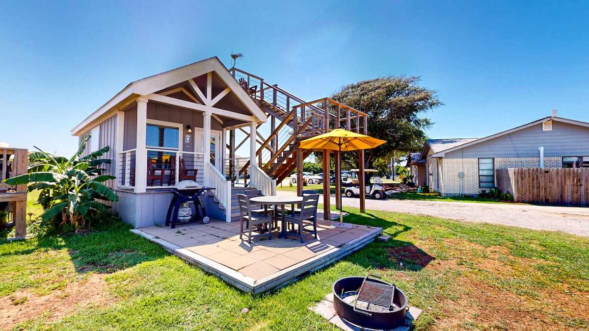 A cabin exterior showcases a spacious patio area with a table and chairs under a yellow umbrella. A grill is positioned nearby, and a fire pit is set on the grass. A staircase leads to an upper level, surrounded by lush greenery.