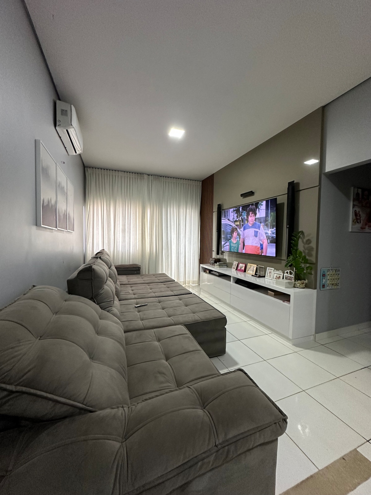 A spacious living area features a light grey sectional sofa with a plush texture, positioned adjacent to a large window draped in sheer curtains. A wall-mounted television is visible, accompanied by a modern media console displaying books and decorative items. Soft lighting enhances the inviting environment.
