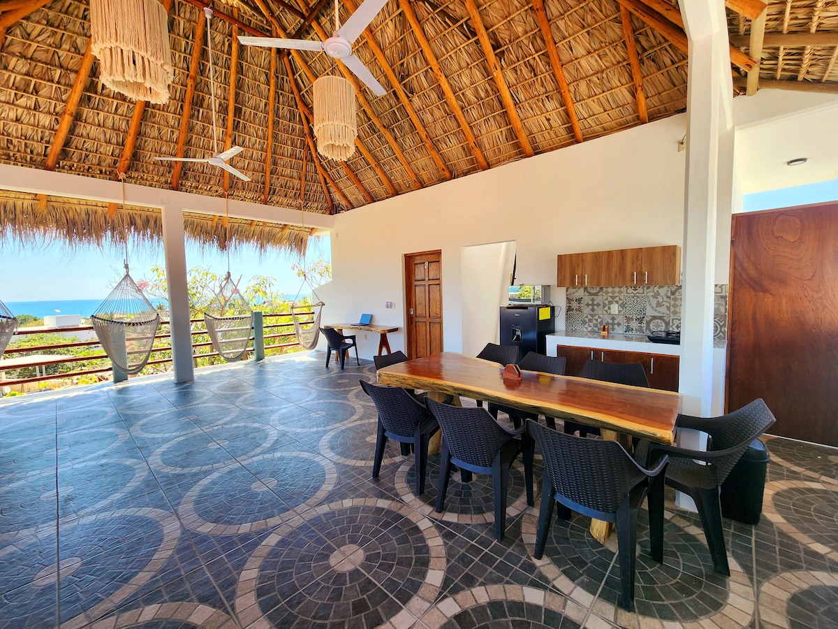 An open terrace area features high thatched ceilings and a spacious layout. A large wooden dining table is surrounded by black chairs. A kitchen area with wooden cabinetry is visible, along with hanging hammocks for relaxation. The ocean view is seen from the terrace.
