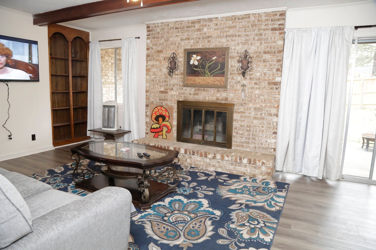 A living area is displayed with a brick fireplace as the focal point. A dark wooden coffee table sits on a richly patterned area rug. Light-colored curtains frame windows, allowing natural light to illuminate the space. A television is positioned on a nearby wall.