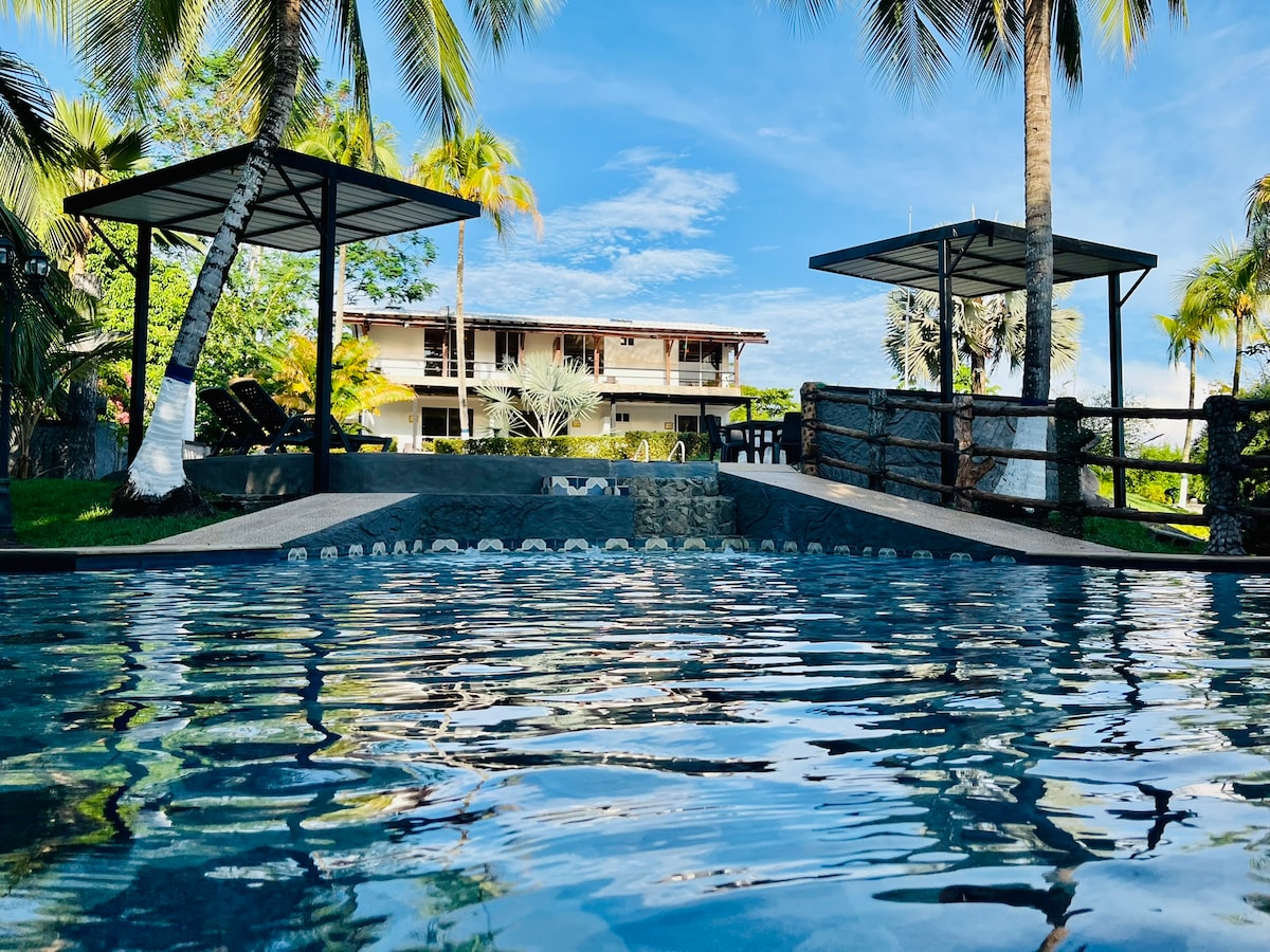 The image captures a tranquil pool reflecting the blue sky and nearby palm trees. Two shaded lounge areas are visible around the pool, with a two-story building situated in the background. Lush greenery surrounds the space, enhancing the serene environment.