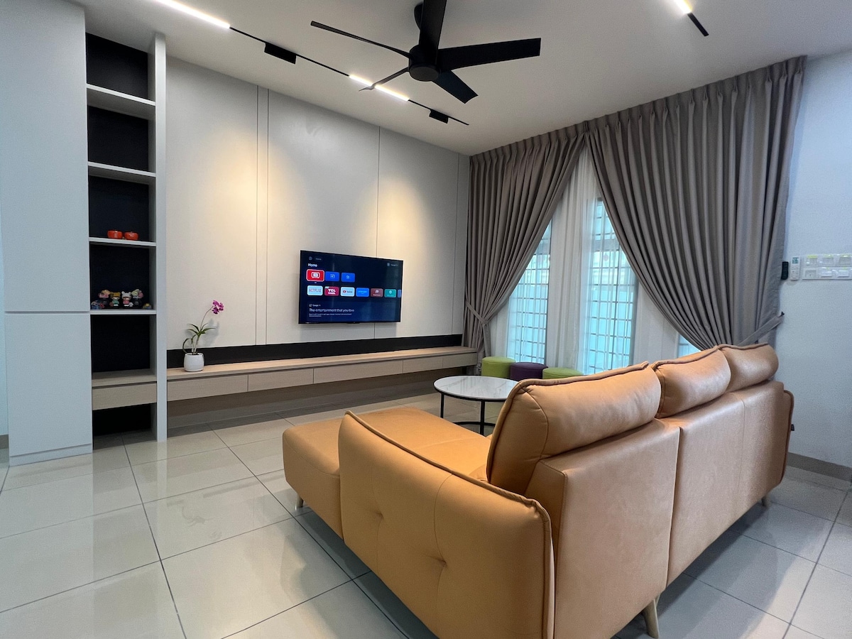 A modern living area is furnished with a leather sofa and a circular coffee table. A wall-mounted television is positioned above a low cabinet, and large windows are dressed with light drapes. A ceiling fan adds to the room's comfort and functionality.