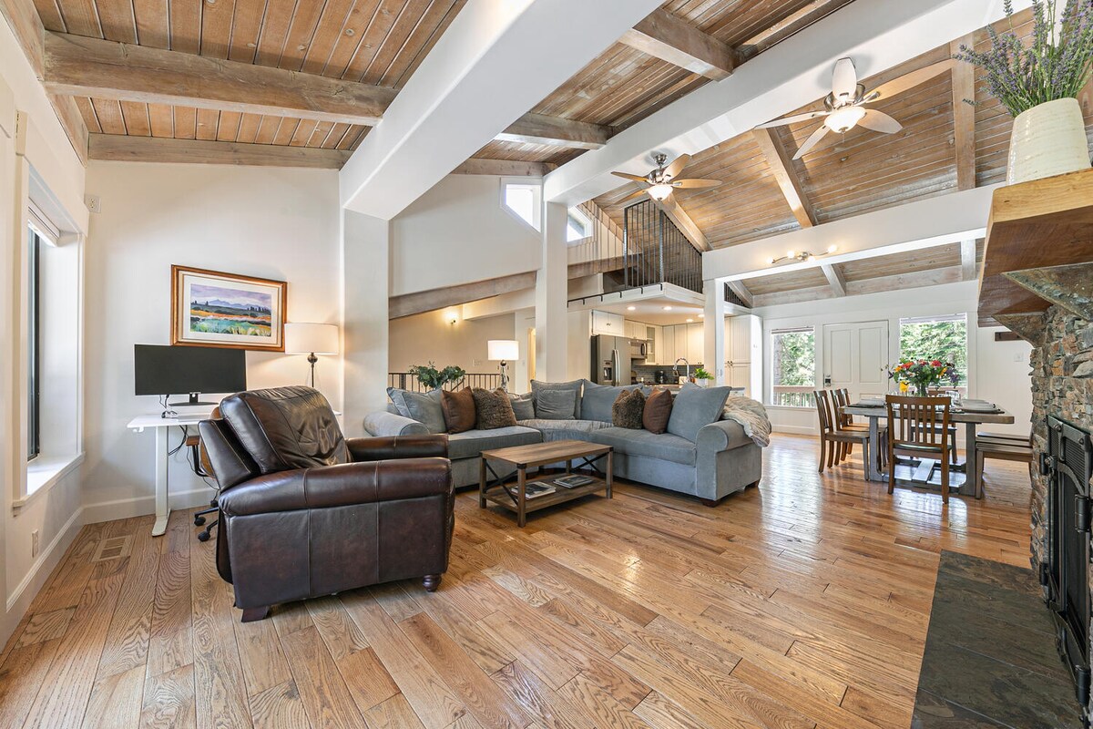 The spacious living area is characterized by high ceilings and abundant natural light. A large sectional sofa is positioned around a coffee table, with a work desk visible to the side. The warm wooden floors add a natural element, complemented by a stone fireplace.