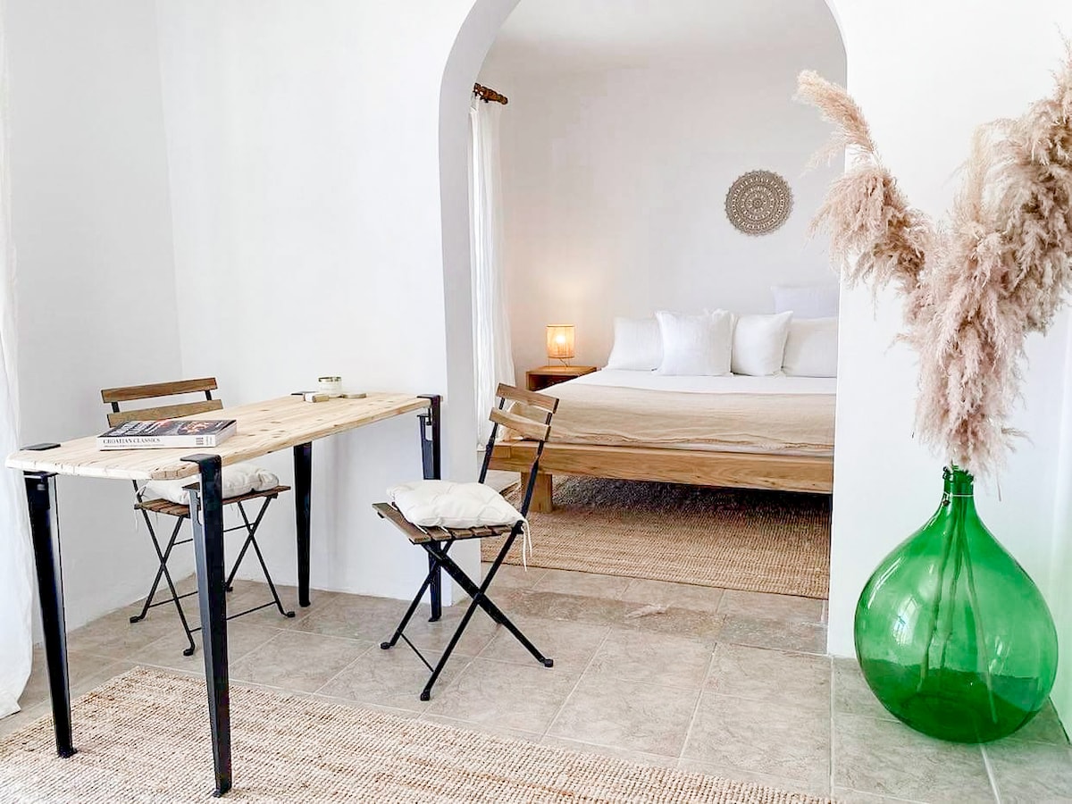 A spacious apartment area features a light wooden table with two foldable chairs beside it. In the background, a king size bed is dressed in white linens, with a warm lamp providing soft lighting. Decorative elements include a large green vase and a textured rug on the floor.