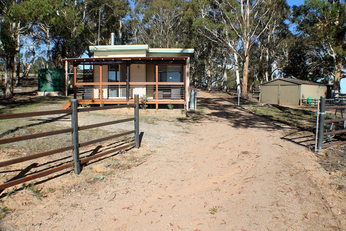 A charming cottage is set amidst a natural landscape, featuring a front porch and surrounded by tall trees. A gravel driveway leads to the entrance, and a shed is visible in the background. A rustic fence frames the property, enhancing the rural appeal.