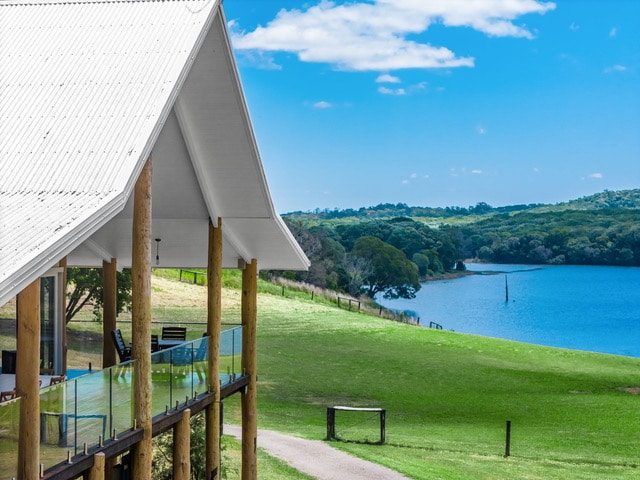 A modern pole home is featured, showcasing a full wrap-around verandah that offers views of Lake Tinaroo and surrounding bushland. The vibrant blue water contrasts with the lush green landscape, while a clear sky adds a serene backdrop.