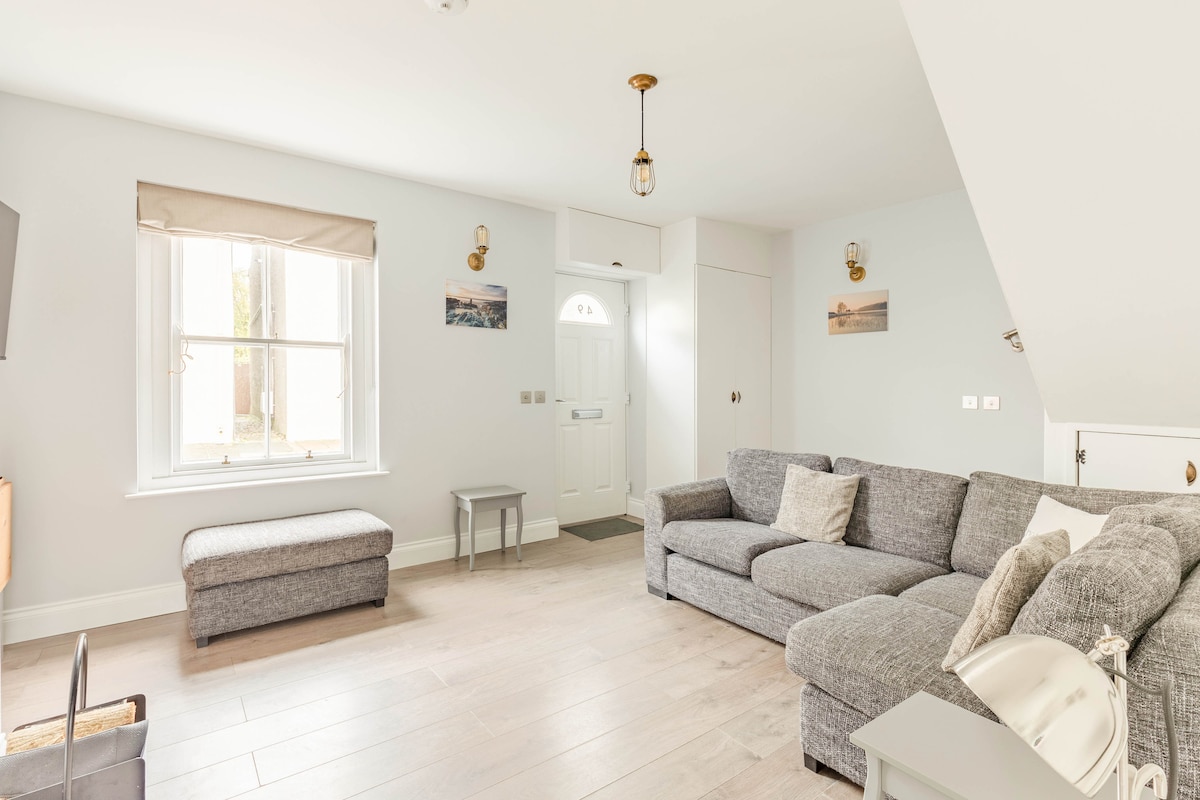 A spacious living area features a large, comfortable grey sectional sofa and a matching ottoman. A light wood floor complements the neutral walls. Natural light enters through the front window, and a door leads outside. Wall decor includes minimalistic photographs.