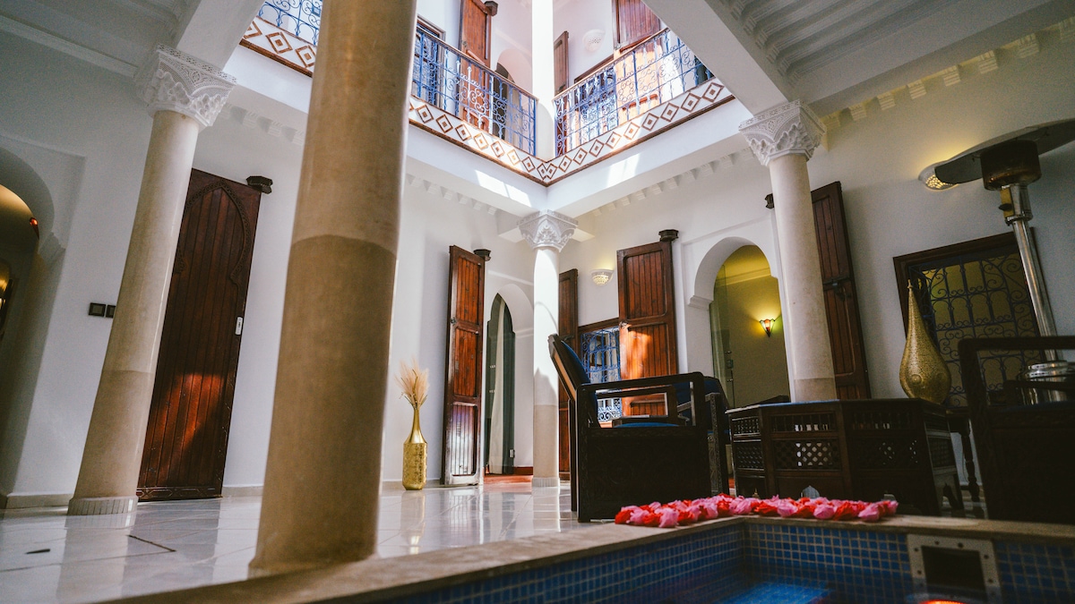 An interior view captures the spacious courtyard featuring decorative columns and a tranquil swimming pool. Natural light filters through the roof, illuminating the elegant seating area adorned with a small table. Traditional Moroccan detailing enhances the cultural ambiance of the space.