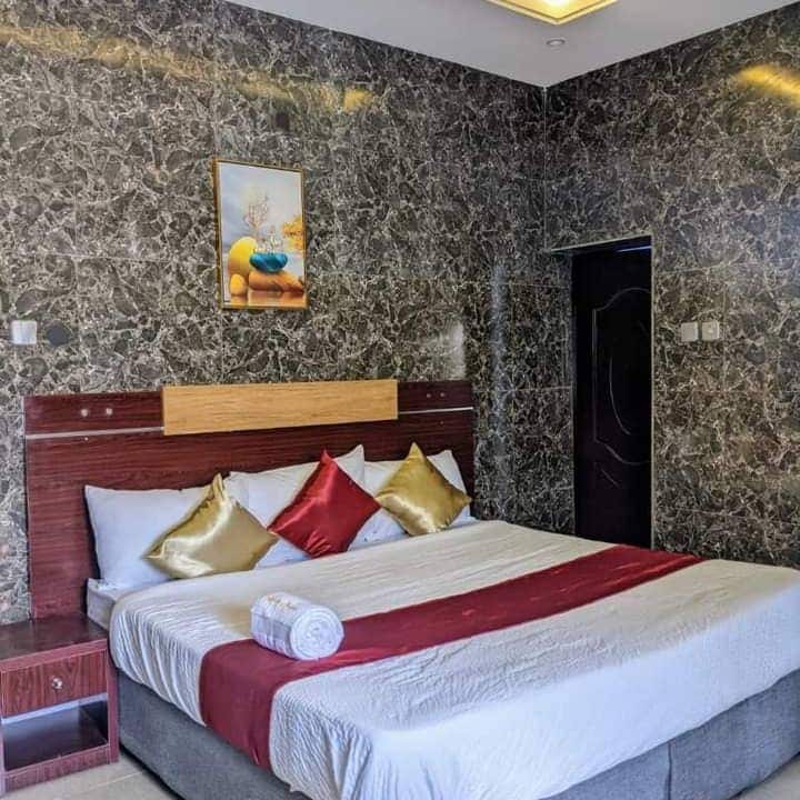 A well-furnished sleeping area features a spacious bed with decorative pillows in shades of red and gold. A neatly rolled towel is placed at the foot of the bed. The wall is adorned with a simple artwork, complementing the marbled texture of the surrounding walls.