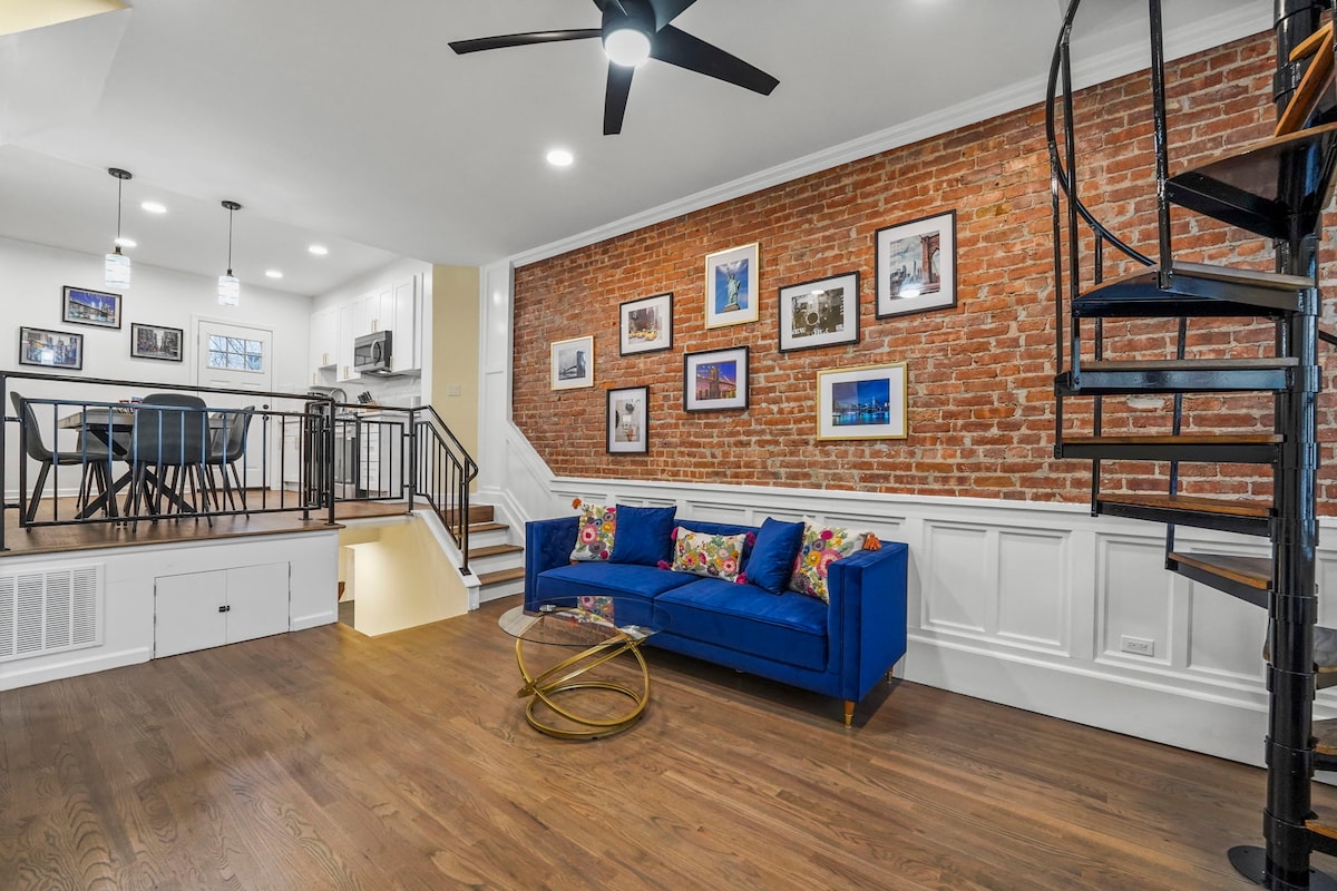 A living area features a stylish blue sofa, accented by colorful pillows, and sits against a brick wall. A spiral staircase is visible, leading to the upper level. The space is illuminated by modern lighting fixtures, and framed photographs are displayed on the walls.