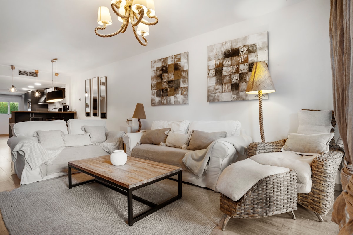 A spacious living room is furnished with a comfortable sectional sofa and an armchair. A wooden coffee table is centered on a neutral rug. Artwork is displayed on the walls, and a stylish chandelier adds ambient light. A glimpse of the kitchen area is visible in the background.