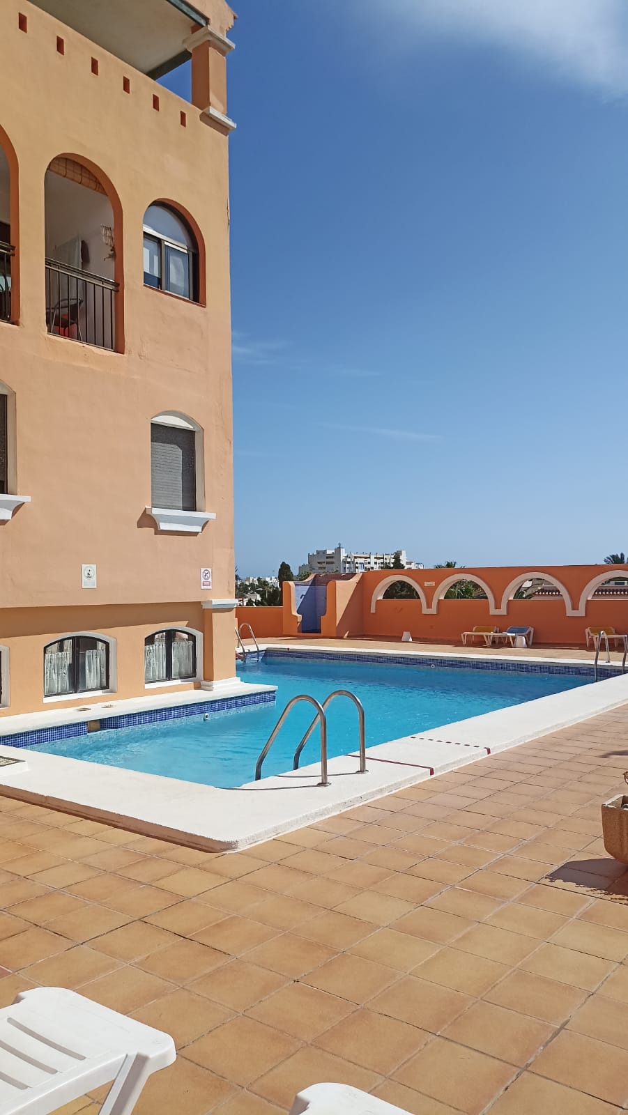 A well-maintained swimming pool is visible, surrounded by a tiled patio. The building features a warm, orange exterior with arched windows. Sun loungers are arranged nearby, providing a relaxing area for guests. Clear blue skies enhance the outdoor setting.