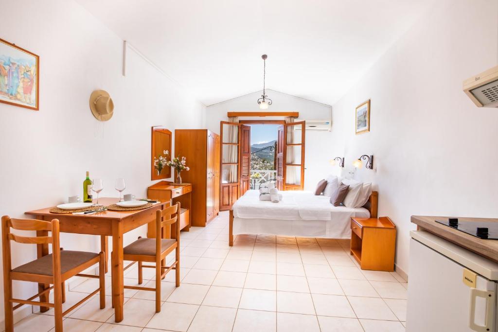 A bright studio space is presented with a spacious bed flanked by two bedside lamps. A wooden dining table with two chairs is arranged next to the window, while a kitchenette and wardrobe are visible in the background. Natural light illuminates the room.