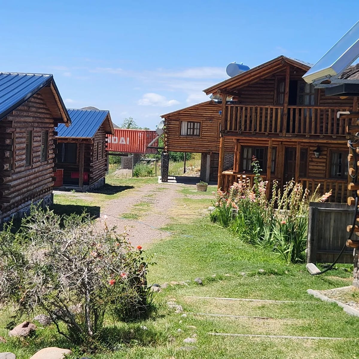 A cluster of rustic wooden cabins is set amidst a lush green landscape. Vibrant flowers line the pathway leading through the yard, while clear blue skies provide a bright backdrop. The cabins feature balconies and varied roof designs, contributing to the inviting outdoor environment.