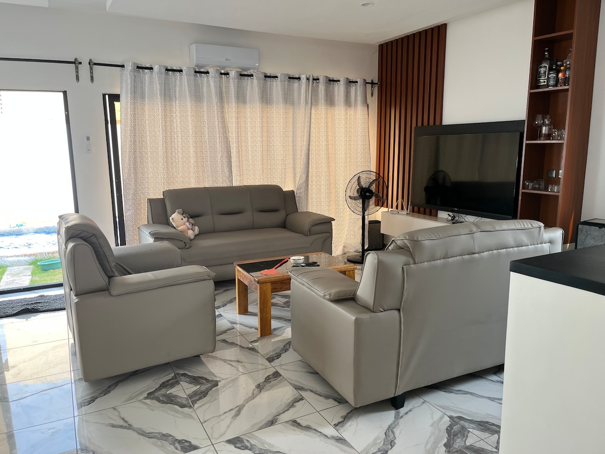 A modern living area is furnished with two light-colored sofas and an armchair, arranged around a wooden coffee table. A large television is mounted on the wall, while sheer curtains provide a soft diffusion of natural light. Decorative elements are placed on the shelves.