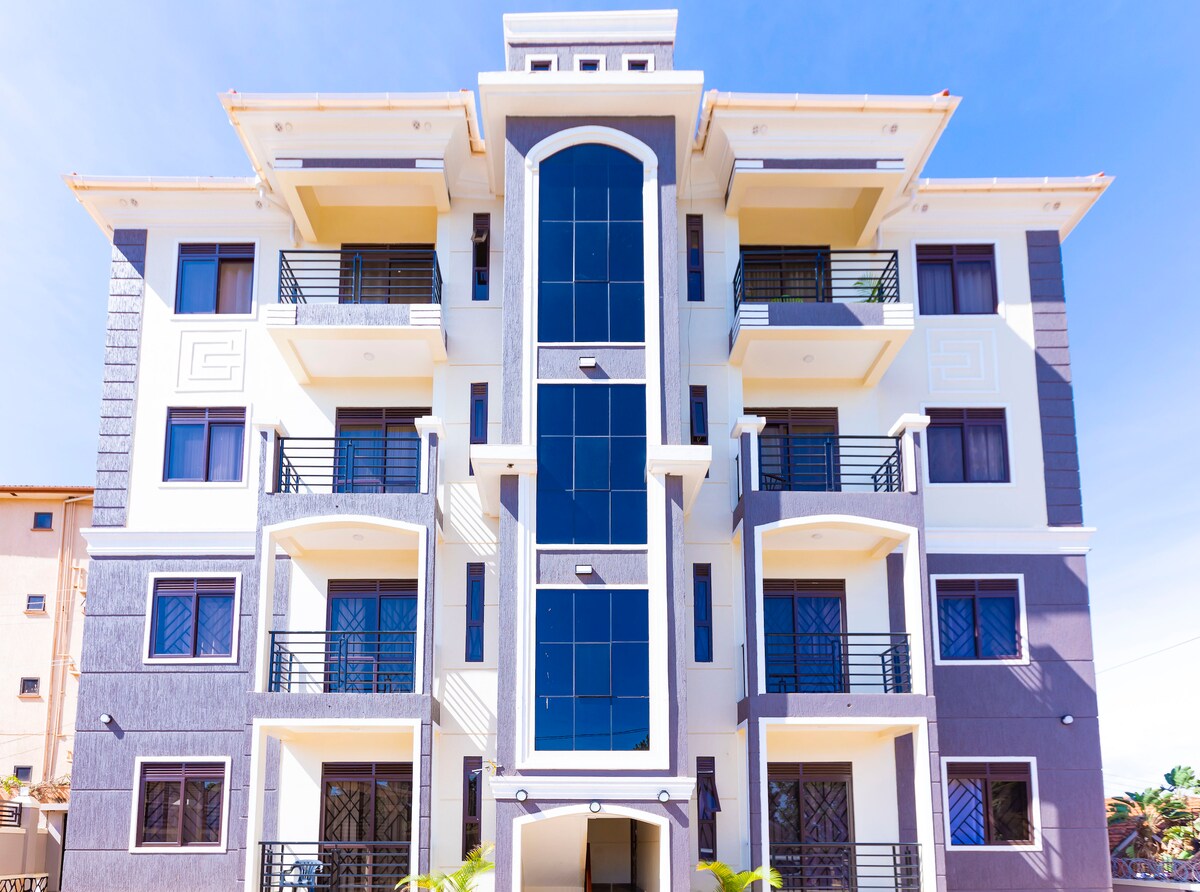 A four-story apartment building is displayed, featuring a modern architectural design. Large windows create a bright facade, while black balconies provide outdoor space for upper units. The building is framed by a clear blue sky, emphasizing its sleek lines and contemporary appearance.